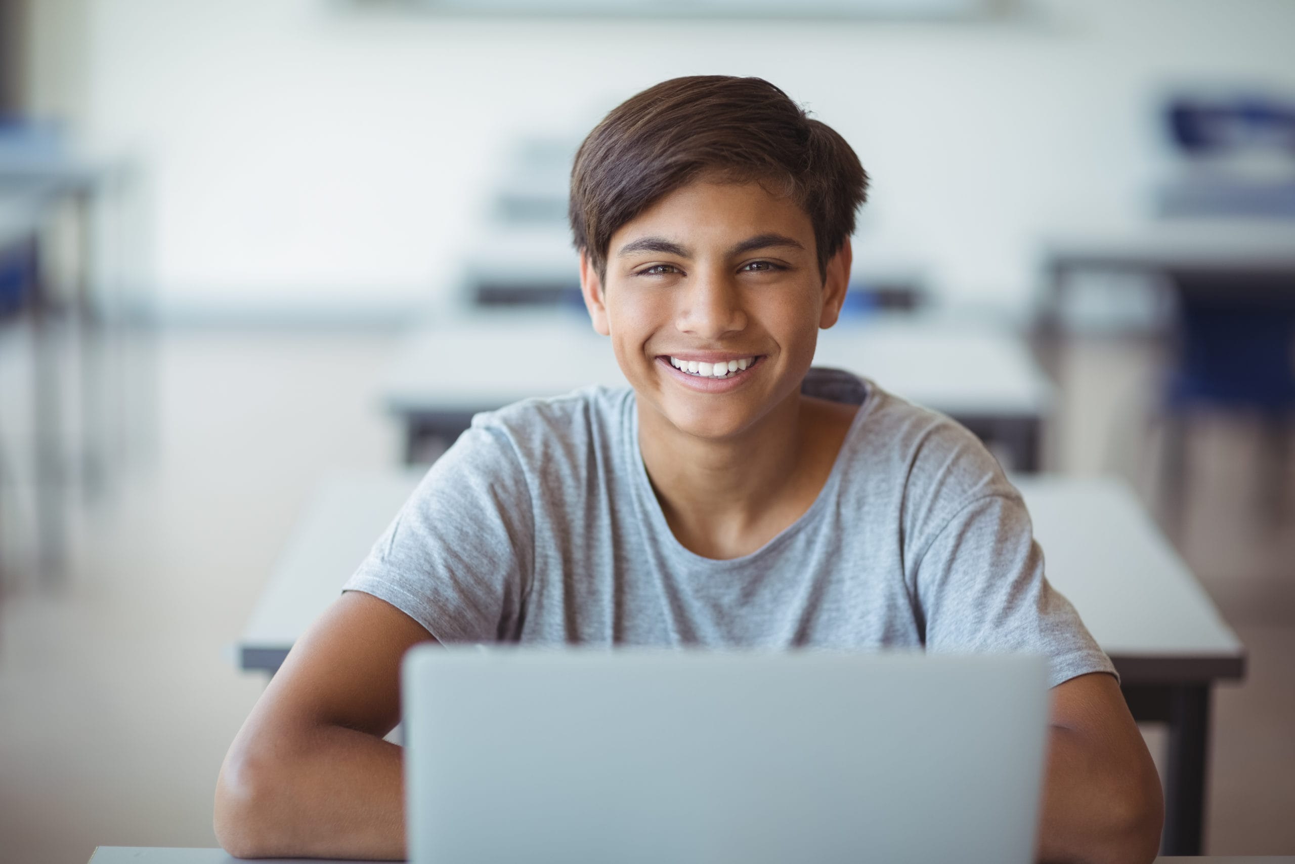 Smiling young male student in a gray t-shirt sitting at a school desk with a laptop, looking directly at the camera in a bright classroom setting.