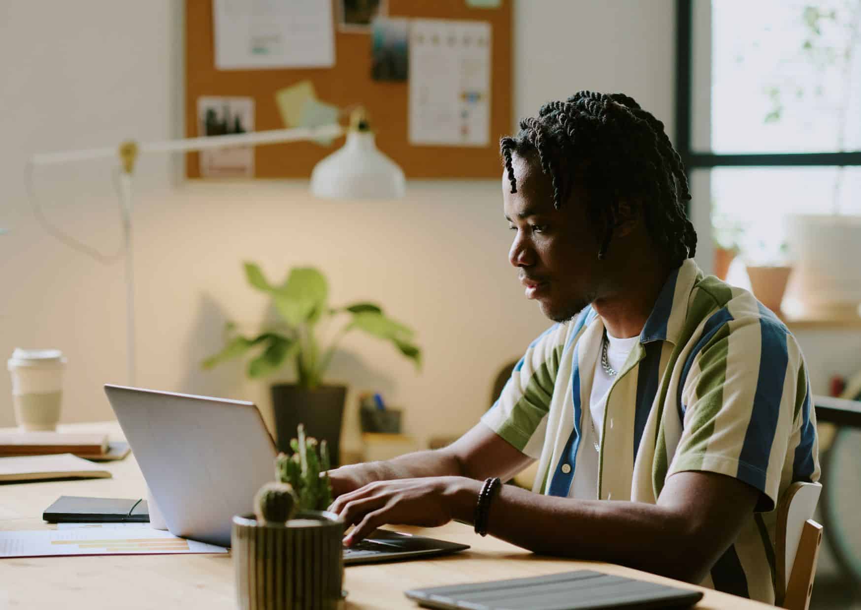 A focused young professional with dreadlocks working on a laptop at a wooden desk in a warm, modern home office.