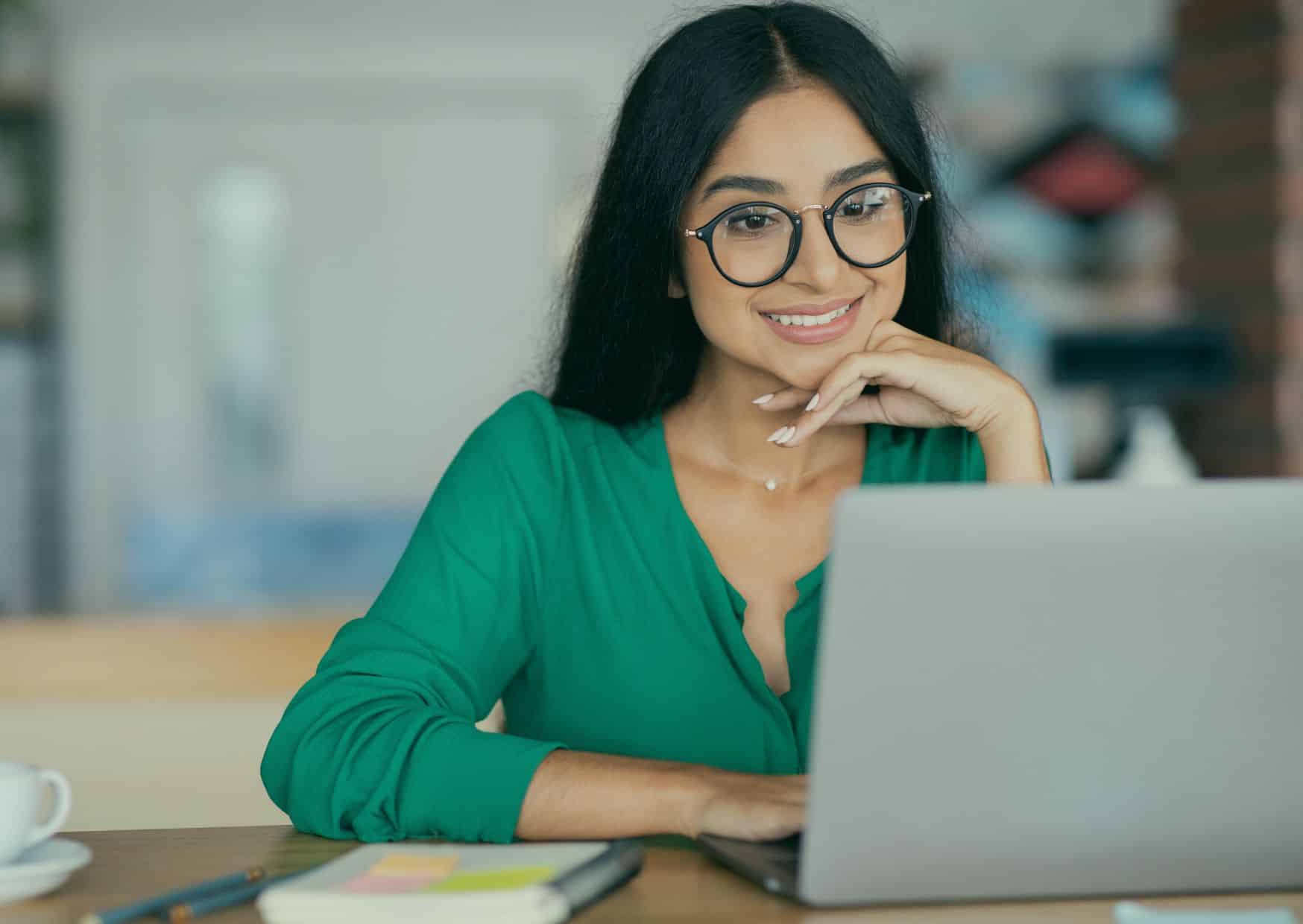 A smiling professional woman with glasses working on a laptop in a bright, modern office or home workspace.